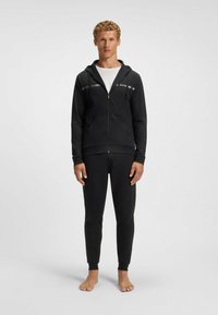 Mustapohjainen vetoketjuhuppari, jossa on heijastavat raidat ja logo, yhdistettynä mustiin slim-fit-housuihin. Materiaalin pinta näyttää sileältä ja joustavalta.