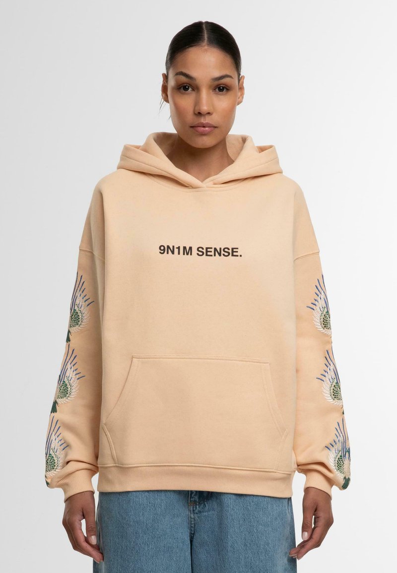 Peach hette med kengurulomme, med trykk av "9N1M SENSE." og broderte blomsterdesign på ermene, ribbestrikkede mansjetter og nederst.