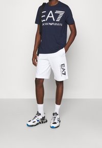Navy cotton t-shirt with white “EA7 Emporio Armani” logo, paired with white shorts featuring a logo print, and black and white sneakers with blue accents.