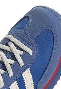 Chaussure de sport bleue en matériau suédé et en mesh, avec des accents à rayures blanches, des lacets texturés et une bordure de semelle rouge.