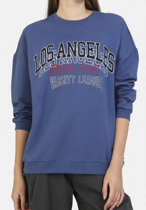 Sweat-shirt bleu avec le texte "Los Angeles Athletic Dept Varsity League" porté par une personne aux cheveux longs et pantalon gris foncé, mains détendues le long du corps.