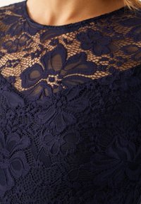 Haut en dentelle navy avec des motifs floraux, une texture superposée et un décolleté transparent, avec une base en tissu lisse contrastant avec le detailing délicat.