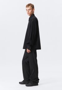 Mustapintainen ylipitkä kauluspaita pitkähihaisilla hihoilla, yhdistettynä vastaaviin löysiin mustapintaisiin housuihin ja teksturoidulla mustalla kengillä.