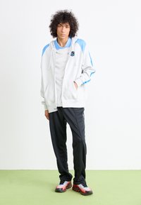 Casaco de capuz branco com detalhes azuis, combinado com uma t-shirt cinza, calças de treino pretas e sapatilhas multicoloridas. Design simples, ajuste casual.