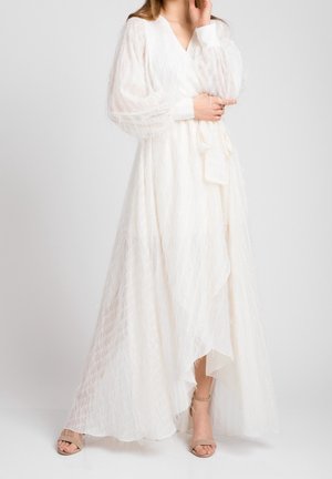 Kobieta ubrana w długą białą sukienkę zapinaną z teksturowanej tkaniny z przejrzystymi bufkami na rękawach, w beżowych sandałkach na obcasie z odkrytymi palcami, stojąca na jednolitym tle.