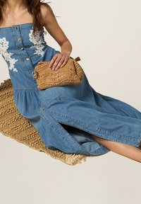 Robe en denim avec broderie florale blanche, boutons sur le devant et fentes sur les côtés. Pochette tressée beige tenue à la main.