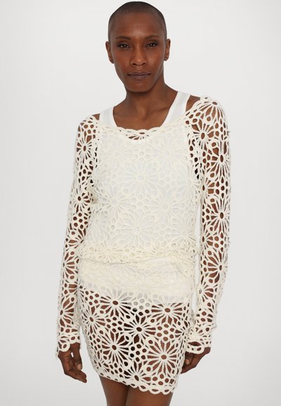 Top croșetat alb cu model floral, mâneci lungi și o croială lejeră, purtat peste un top alb. Textură transparentă cu detalii intricate.