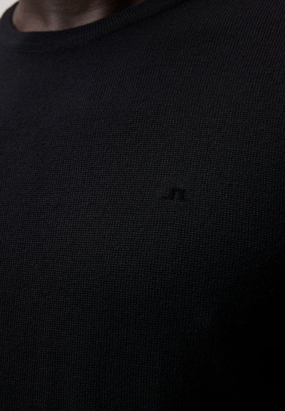 Juodas megztinis su apvaliu iškirpimu, su subtiliu iškilniu logotipu ant krūtinės. Audinys pasižymi smulkia, vienoda audimo struktūra.