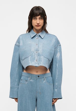 Moteris dėvi šviesiai mėlyną trumpą džinsinę striukę ir atitinkančius aukšto liemens džinsus su blizgučių detalėmis ant striukės ir kišenių.