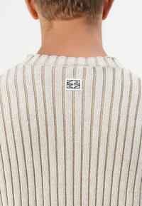 Pull en tricot beige et crème avec des rayures verticales. Présente un col côtelé et une petite étiquette carrée à l'arrière, près de l'encolure.