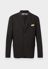 Μαύρο καλοραμμένο σακάκι με δύο κουμπιά μπροστά, τσέπες με καπάκι και μια χρυσή ετικέτα "Versace Jeans Couture" στην τσέπη του στήθους.