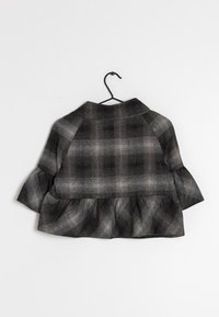 Kort svart och grå rutiga jacka med volangkant som hänger på en svart trådhängare mot en vit bakgrund.