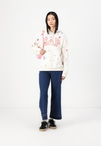 Sudadera crema con estampado floral y lettering rojo "GAP", combinada con jeans de pierna ancha de color azul oscuro y zapatillas negras. Acento de bolso rosa.