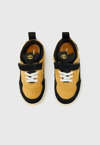 Brown suede sneakers with black accents, featuring white laces, a velcro strap, and a logo on the tongue, designed for comfort and support.
