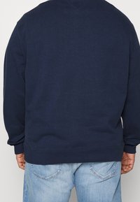 Felpa blu navy con polsini e orlo a coste, realizzata in tessuto morbido, caratterizzata da una vestibilità comoda e un design del retro senza cuciture. Indossata con jeans blu chiaro.