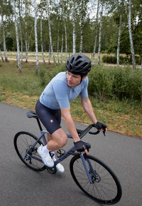 Cycliste portant un maillot bleu clair et un short noir, montant un vélo gris sur un chemin pavé entouré de bouleaux.