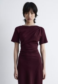 Burgunderrotes Kleid mit kurzen Ärmeln und drapiertem Design, aus glattem Stoff und mit taillierter Silhouette. Eine einfache Silberkette wird getragen.