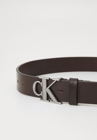 Hnedý kožený opasok s kovovou sponou v striebornej farbe s logom "CK", so stručným prešívaním po celej dĺžke a hladkou textúrou.
