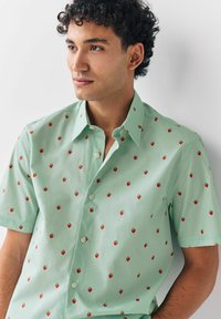 Hombre con cabello rizado que lleva una camisa de botones de manga corta de color verde claro con un estampado de pequeñas fresas, de pie contra una pared blanca.