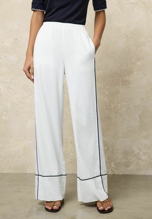 Nő fehér, fekete szegélyű, bőszárú nadrágban, zsebekkel és barna nyitott orrú szandálban áll egy texturált bézs fal előtt.