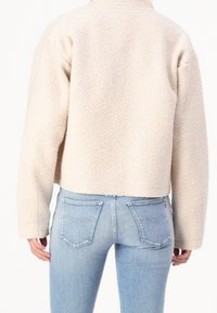 Krämfärgad, texturerad fleecejacka med en kort design och hög krage, bärs över ljusblå denimjeans med bakfickorna synliga.