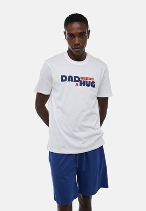 T-shirt blanc à manches courtes avec "DAD NEEDS A HUG" imprimé en bleu et rouge. Associé à un short bleu. Matière en coton.