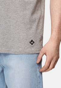 Grå bomulds t-shirt med korte ærmer, der har et sort diamantformet logo på sømmen og kontraststing. Båret med lyseblå jeans.