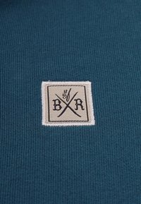 Teal strikket stof med en kvadratisk hvid lap, der har bogstaverne "B/R" og krydsede linjer. Teksturen er glat med et ribbet mønster.