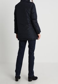 Zwarte pufferjas met een hoge kraag, lange mouwen en een zoom die onder de heupen eindigt, gecombineerd met donkere denim jeans en met hakken enkellaarsjes.