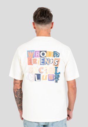 Hombre con el pelo corto y tatuajes en el brazo izquierdo, que lleva una camiseta blanca con letras de colores en bloque y un pequeño dibujo azul de un osito de peluche en la espalda.