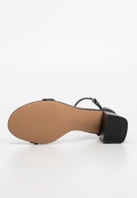 Sandale noire avec une semelle beige et un petit talon bloc, vue du dessous sur fond blanc.