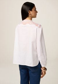 Blusa bianca con maniche lunghe e dettagli in pizzo sul retro. Orlo arrotondato e leggero volume sulle spalle. Indossata con jeans blu.