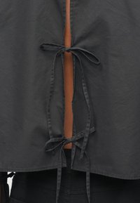 Top scurt negru realizat dintr-un material ușor, cu o închidere la spate cu șnur și un design decupat care expune o secțiune contrastantă de culoare maro.