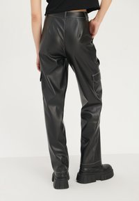 Pantalons cargo en simili cuir noir avec une texture lisse, dotés de poches latérales et de coutures blanches contrastantes, associés à des chaussures noires chunky.