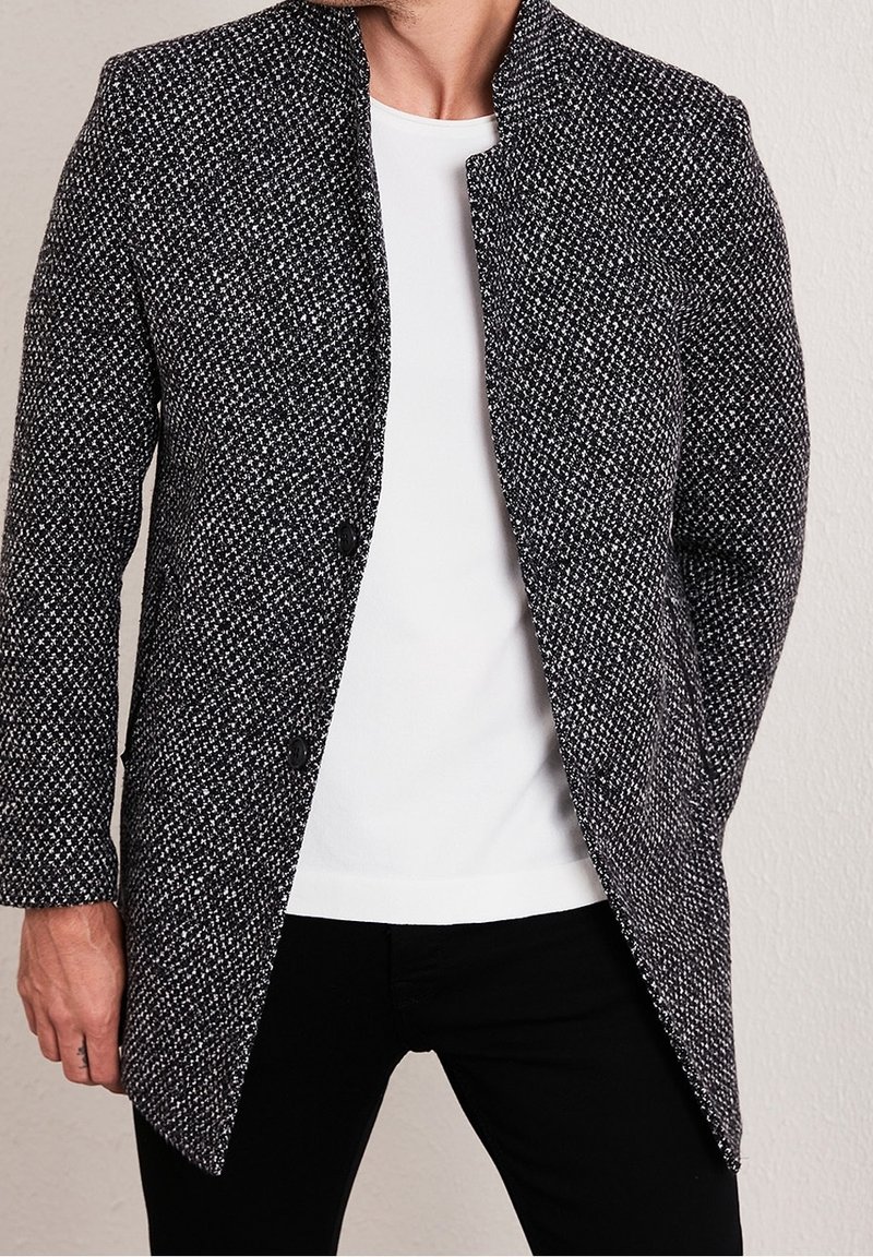 Kabát z vlněné směsi s texturovaným černo-bílým houndstooth vzorem, mandarin límcem a dvouřadým zapínáním na knoflíky, kombinovaný s bílou košilí.