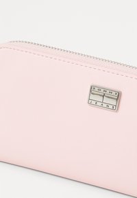 Portafoglio sintetico rosa con una texture liscia, chiusura con zip e dettagli in argento, caratterizzato da una placca rettangolare con il logo "TOMMY JEANS".