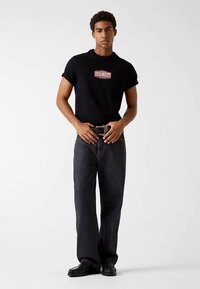 Zwart T-shirt met korte mouwen met een rood-wit "Guess Jeans" logo, gecombineerd met een donkere spijkerbroek en zwarte schoenen, staand tegen een effen achtergrond.