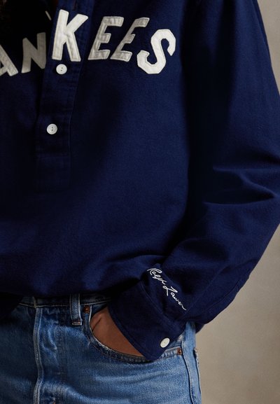 Camisa de manga longa azul-marinho com um logótipos "Yankees" branco no peito, colarinho abotoado e detalhe bordado na manga, combinada com denim.