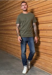 Olivově zelené bavlněné tričko, přiléhavý střih, krátké rukávy. Nosené s modrými džíny a bílými teniskami. Viditelná tetovaná ruka. Dřevěná stěna v pozadí.