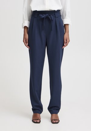 Mujer que lleva pantalones de talle alto azul marino con cinturón de lazo, combinados con blusa blanca de manga larga y tacones abiertos negros.