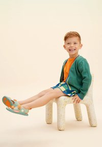 Un bambino è seduto su una sedia bianca maculata. Indossa un cardigan verde, una maglietta arancione, pantaloni corti colorati e sandali blu con dettagli arancioni.