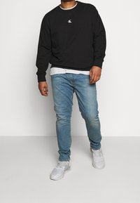 Black long-sleeve sweatshirt with white logo, layered over a white shirt, paired with light blue denim jeans and white sneakers.