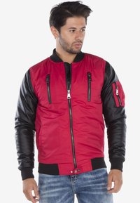 Rode en zwarte bomberjack met een ritssluiting aan de voorkant, twee borstzakken en een zak op de linkermouw. Voorzien van ribbels aan de manchetten en de zoom.
