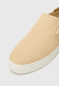 Zapato de lona beige sin cordones, hecho de tela texturizada, con punta redondeada, paneles elásticos a los lados y suela de goma blanca.
