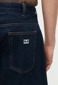 Κοντινό πλάνο πίσω τσέπης σκουρόχρωμης μπλε τζιν φούστας με πορτοκαλί ραφές και μικρό ασπρόμαυρο πατς που δείχνει δύο στυλιζαρισμένα ζώα.