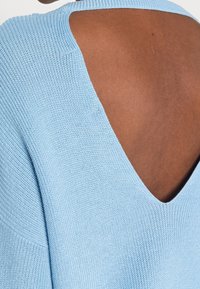 Jasnoniebieski sweter z dzianiny, z głębokim dekoltem w kształcie V z tyłu, posiadający teksturowany materiał i szerokie rękawy, podkreślający detal otwartego pleców.