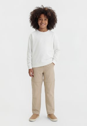 Kind met krullend haar dat lacht, gekleed in een wit sweatshirt, beige broek met een korte bovenlaag en beige instapschoenen, staand met één hand in de zak.