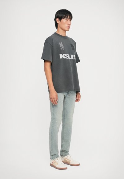 Grijze oversized t-shirt met KSUB-logo, gecombineerd met lichtblauwe distressed jeans en witte sneakers met tan accenten.