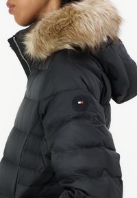 Crni puffer kaput s kapuljačom obrubljenom krznom, kvadratičastom teksturom, označenom zakrpom na rukavu i detaljima od srebrnog zatvarača.