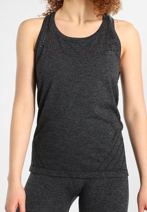 Kobieta w ciemnoszarym bezrękawnym sportowym topie i dopasowanych legginsach, stojąca z rękami swobodnie opuszczonymi wzdłuż ciała na jasnym tle.